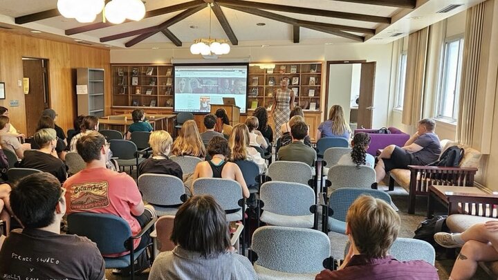 Classroom setting with attendees facing a presenter and screen in a wood-paneled room.