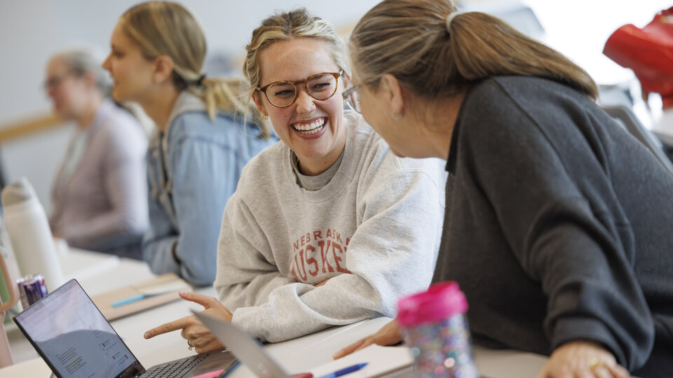 Two teachers smile at each other while pointing to a computer during the Summer Institute